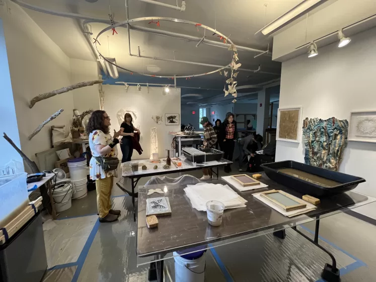 A picture of a white walled studio space with table holding a paper making station. People begin entering the room from the background.