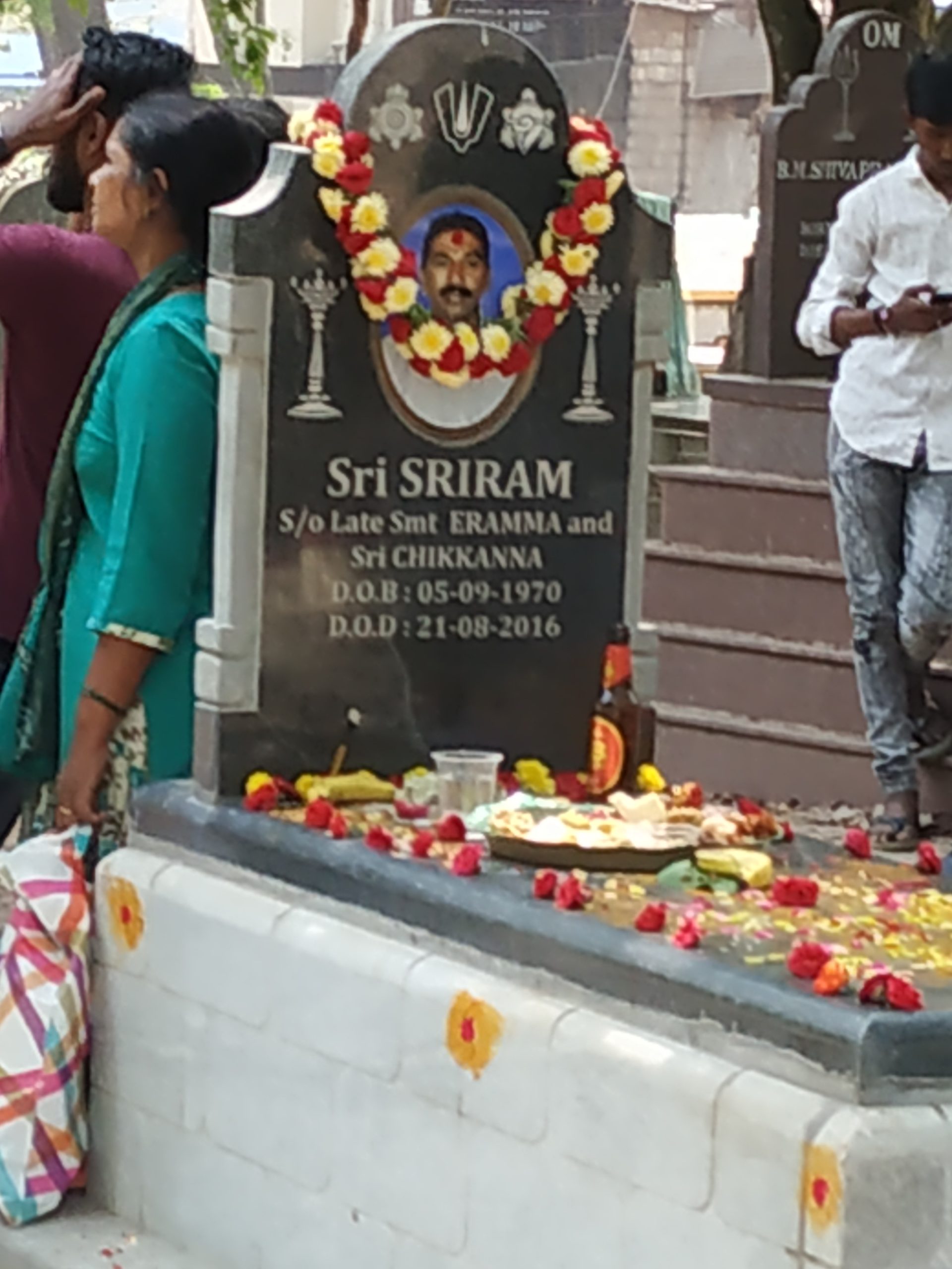 A group of people standing around a grave covered in flowers, food, and glasses of beer