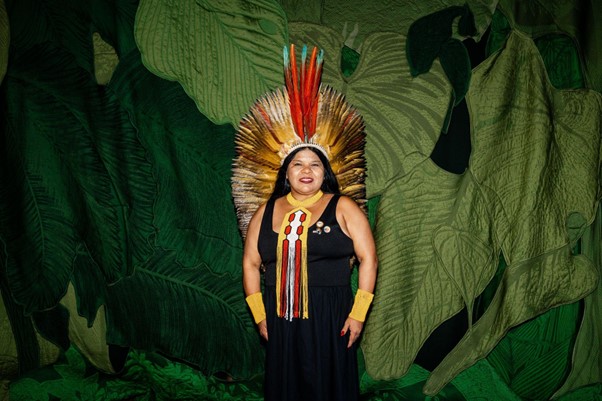 A person standing in traditional headdress and clothes in front of a leafy green background