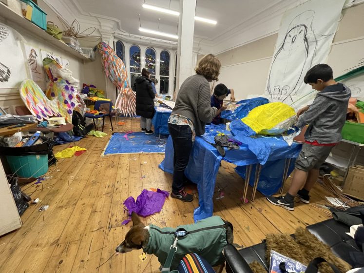 A picture of people and a dog in a craft workshop with lots of cloth, props, and colorful textiles around the room