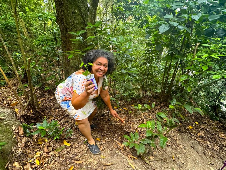 A picture of a smiling woman holding a small plant in a forest